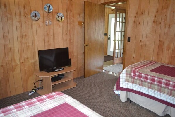 A wooden-paneled room with a television on a wooden stand, two beds with plaid bedding, and a door leading to another room.