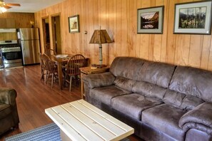 A cozy living room with a sofa, a dining table with chairs, a lamp, and a kitchen area with a refrigerator and stove.
