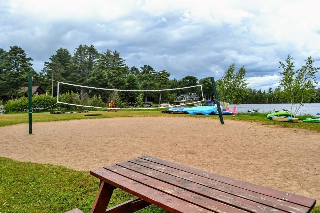 A sandy volleyball court with a wooden bench in the foreground, surrounded by trees and a lake.