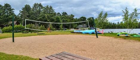 A sandy volleyball court with a wooden bench in the foreground, surrounded by trees and a lake.