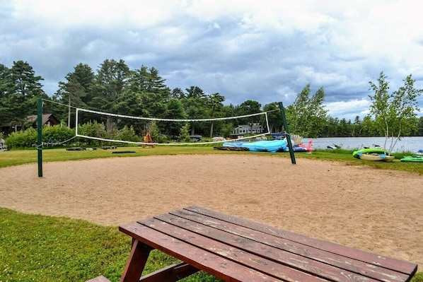 Un terrain de volleyball de plage, avec un banc en bois au premier plan, entouré d’arbres et bordé par un lac.