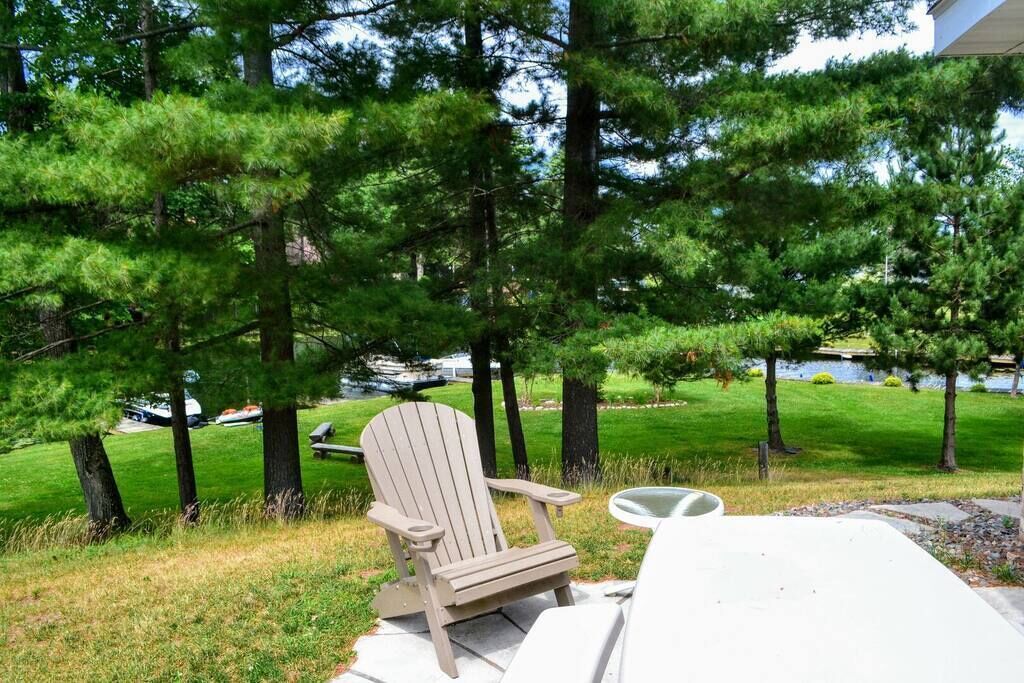 A wooden Adirondack chair with a white cushion on a concrete patio, overlooking a grassy area with trees and a body of water in the background.