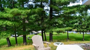 A wooden Adirondack chair with a white cushion on a concrete patio, overlooking a grassy area with trees and a body of water in the background.