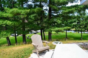 A wooden Adirondack chair with a white cushion on a concrete patio, overlooking a grassy area with trees and a body of water in the background. - Nitschke's Resort Cabin #15 (Minocqua)