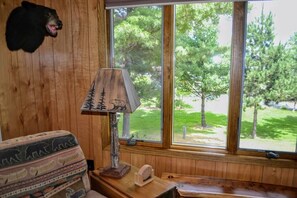 A room with a wooden wall, a bear head decoration, a lamp with a tree design, a patterned cushion, and a window with a view of trees.
