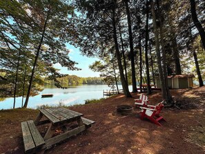 Un terrain de camping au bord du lac, équipé d’une table de pique-nique, de chaises Adirondack rouges et d’un petit abri.