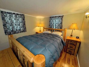 Un dormitorio con estructura de cama de madera, colcha azul, dos mesitas de noche con lámparas y cortinas estampadas.