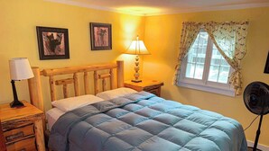 Une chambre à coucher avec une tête de lit en bois, un lit recouvert d’une courtepointe bleue, une table de chevet avec une lampe, une commode de chevet avec des tiroirs, une fenêtre avec des rideaux et un ventilateur sur pied.