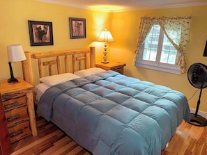 Une chambre à coucher avec une tête de lit en bois, un lit recouvert d’une courtepointe bleue, une table de chevet avec une lampe, une commode de chevet avec des tiroirs, une fenêtre avec des rideaux et un ventilateur sur pied.