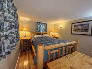 A bedroom with a wooden bed, two bedside tables, a wooden nightstand, a wooden table, a wall-mounted lamp, and a framed picture on the wall.