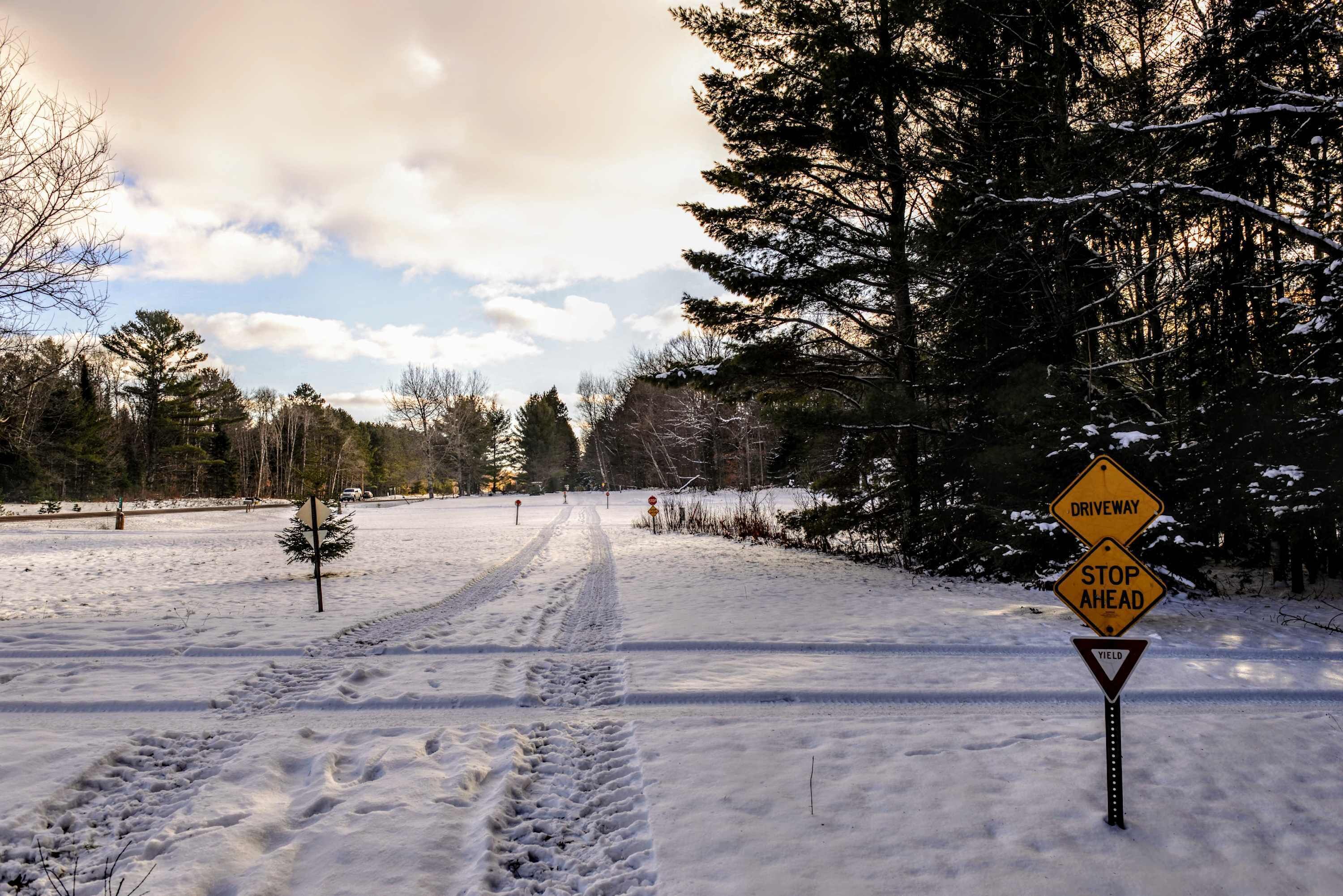 A snow-covered road with a 'Driveway' and 'Stop Ahead' sign, surrounded by trees and a clear sky.