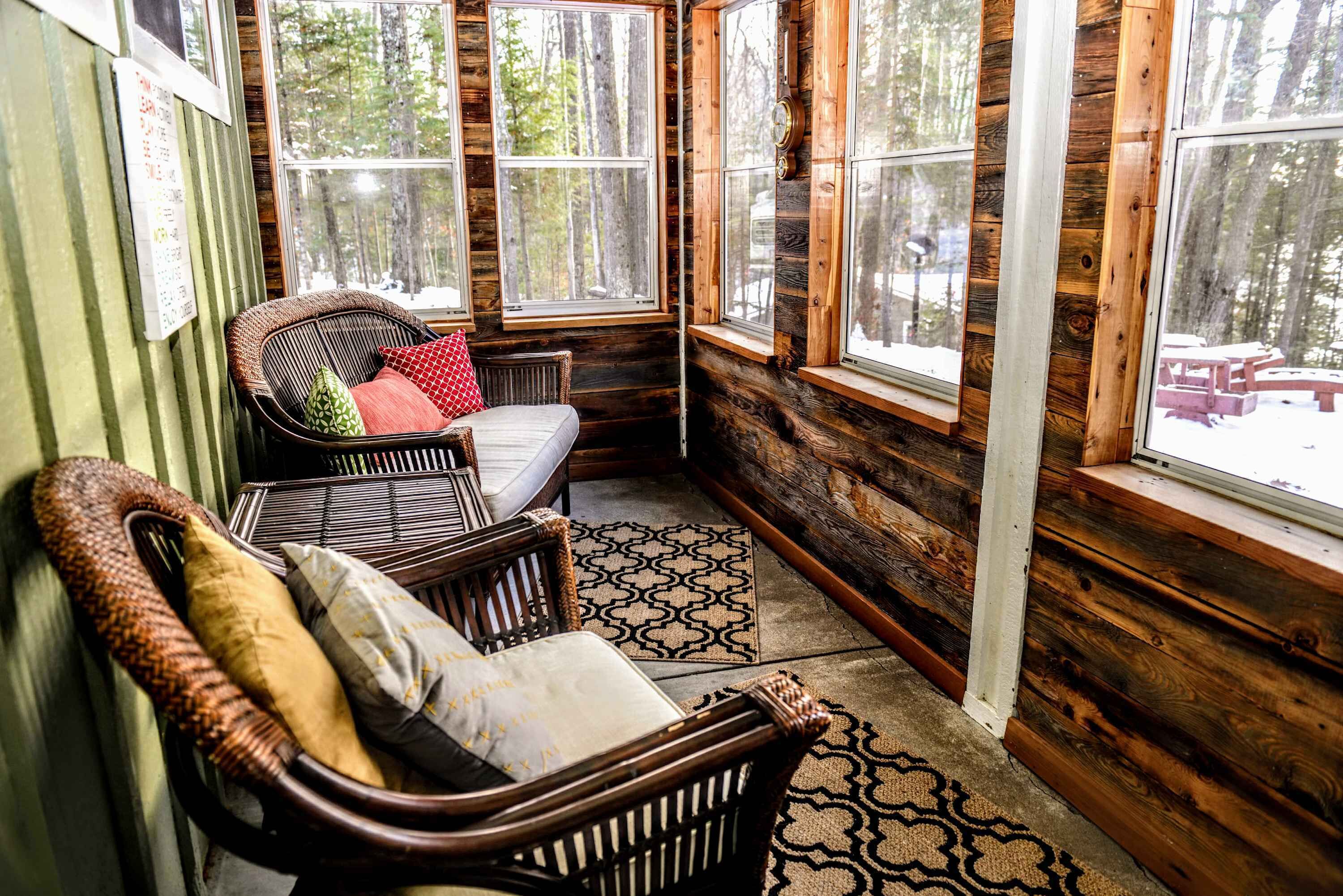 A cozy cabin interior with wooden walls, a wicker chair with cushions, a small bed, and large windows offering a view of the forest.