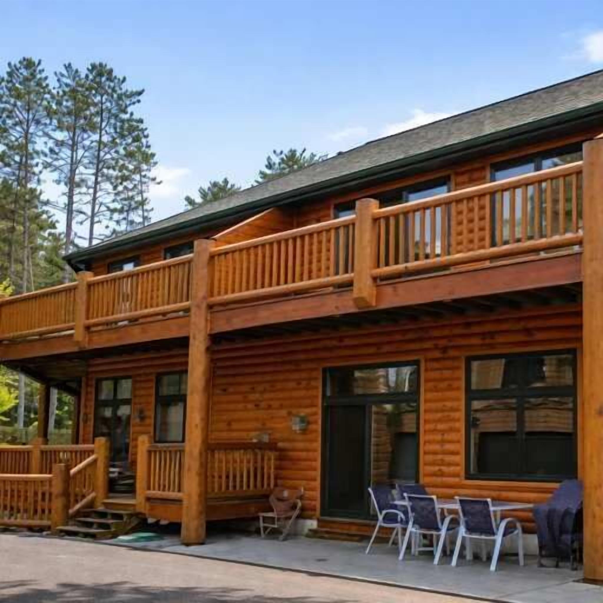 A two-story log cabin with a balcony, outdoor seating, and a view of the surrounding forest.