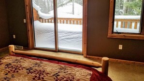A bedroom with a large window, a bed with a floral bedspread, and a view of a snowy deck outside. - Minocqua Lake Condo B10 (Minocqua)