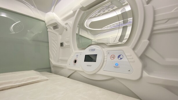 Quarto moderno e bem iluminado, com painel de controle para uma máquina de massagem, cama com cobertura estampada e divisória de vidro.
