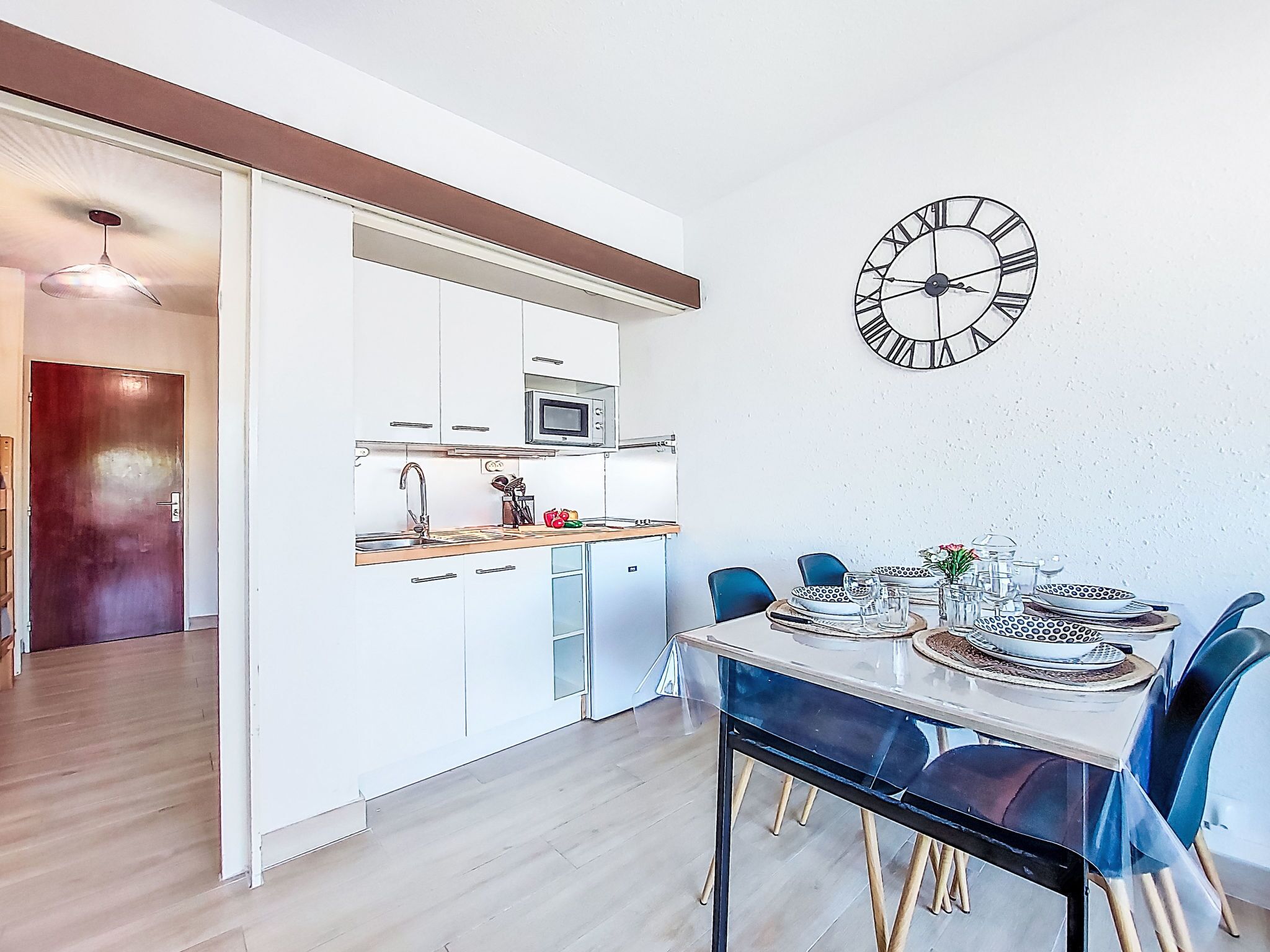 A modern kitchen with white cabinets, a microwave, and a sink. There is a dining area with a table set for two and blue chairs. A wall clock is visible.