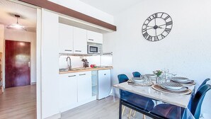 A modern kitchen with white cabinets, a microwave, and a sink. There is a dining area with a table set for two and blue chairs. A wall clock is visible.