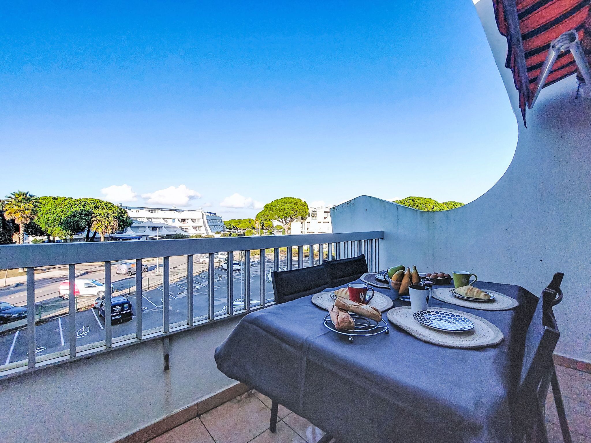 Un balcone con tavolo apparecchiato per mangiare, affacciato su una strada con auto parcheggiate e palazzi.