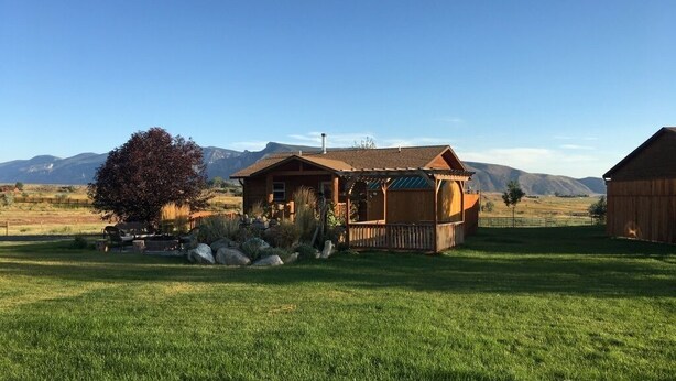 Ein Holzhaus mit Veranda, umgeben von einer grünen Wiese und Bergen im Hintergrund.