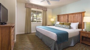 A hotel room with a bed, a TV, a wooden nightstand, a lamp, a ceiling fan, and a window with curtains.