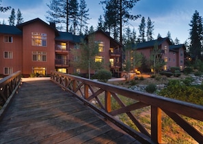 A wooden bridge leading to a lodge with multiple floors and balconies, surrounded by trees.
