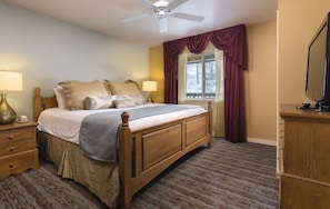 A bedroom with a wooden bed, two bedside tables with lamps, a ceiling fan, a window with curtains, and a television on a wooden cabinet.