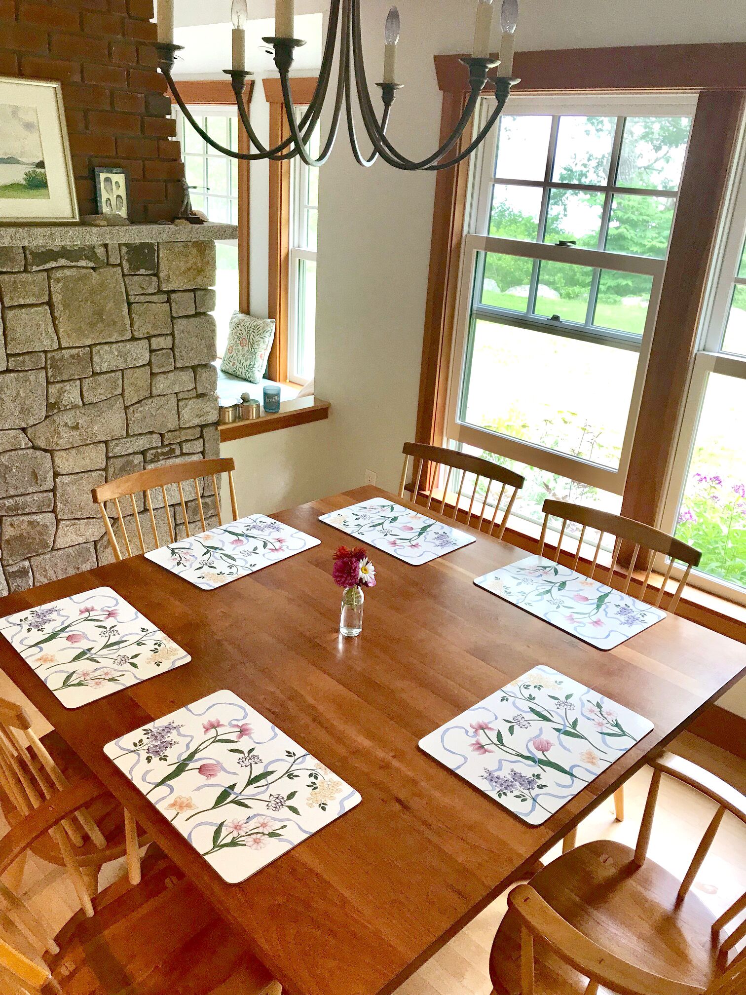 Una mesa de comedor con individuales estampados con flores, sillas de madera, pared de piedra, candelabro y ventana con vistas a follaje.