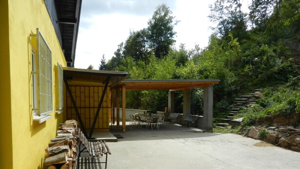 Ein überdachter Außenbereich mit Tischen und Stühlen, einer Holzpergola und einem Holzstoß an einem gelben Gebäude.