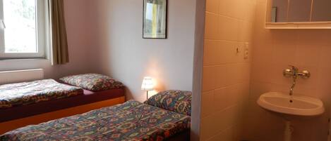 Una habitación pequeña con dos camas, una ventana, un lavamanos instalado en la pared y un cuadro enmarcado.