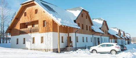 Fila de edificios de estilo chalet de madera con techos nevados, un coche blanco estacionado al frente y un cielo azul despejado.