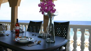 A table set for dining with a bottle of wine, plates, cutlery, and a vase with purple flowers. The table is outdoors with a view of the sea. - Sea-view Apartment in Mambo Beach Near Seaaquarium (Willemstad)