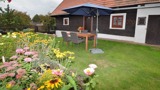 A wooden house with a tiled roof, a garden with various flowers, and a table with chairs under a blue umbrella. - Holiday Home in Forest With Private Pool (Petrohrad)