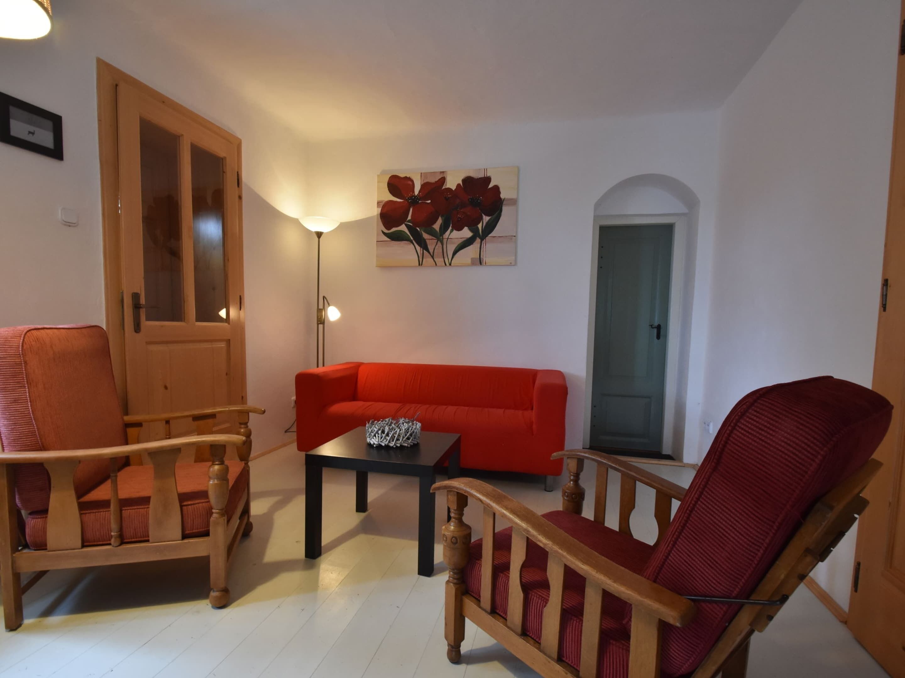 A living room with a red sofa, wooden armchairs, a coffee table, and a painting of red flowers.
