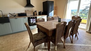 Un coin repas avec une table en bois et des chaises en osier, une cuisine avec des meubles bleu clair, une hotte noire, et une vue sur l’extérieur grâce à une grande fenêtre.