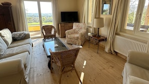 Ein Wohnzimmer mit einer Couch, einem Sessel, einem Tisch, einem Fernseher und Blick ins Freie.