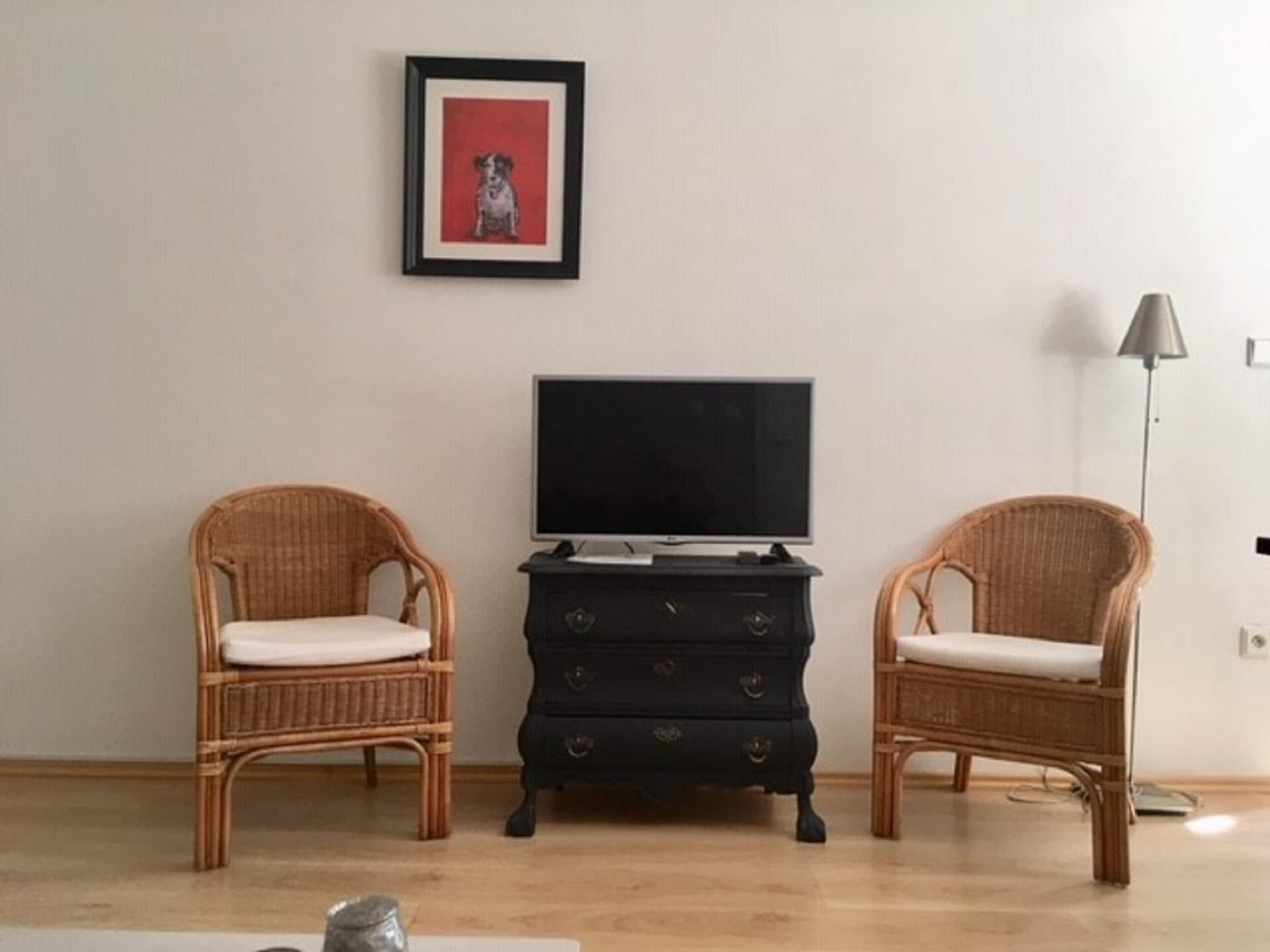 A room with two wicker chairs, a black dresser with drawers, a television on top, and a framed picture on the wall.