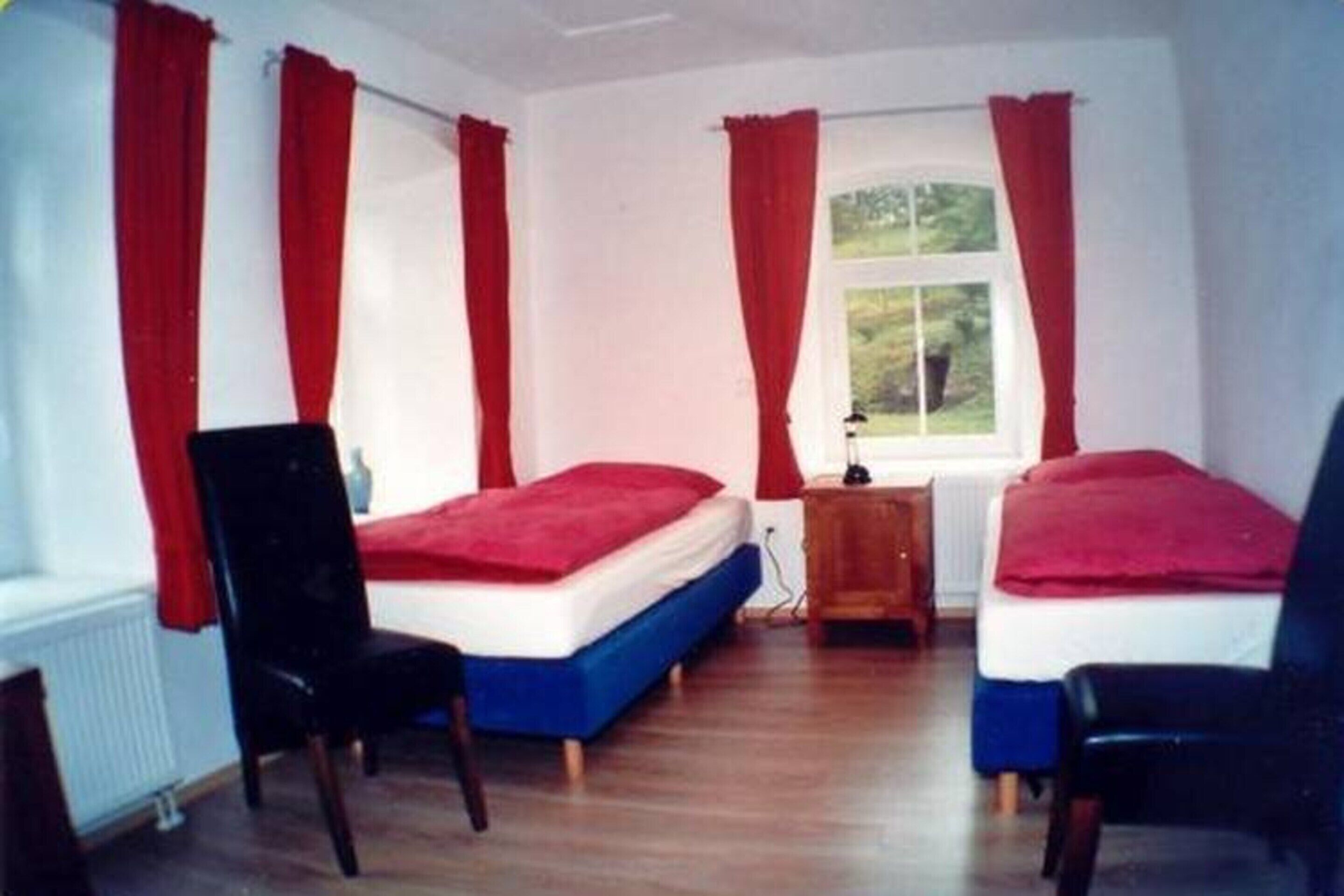 Ein Zimmer mit zwei Einzelbetten, jeweils mit einer roten Decke, Holzstühlen und einem Fenster mit roten Vorhängen.