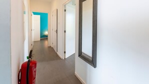 Een gang met witte wanden, vloerbedekking van grijze tegels en deuren die naar andere ruimtes leiden. Een rode brandblusser hangt aan de muur.