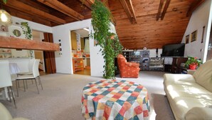 Un salon chaleureux avec un pouf à carreaux, un canapé et un téléviseur. Le plafond est en bois et une plante y est suspendue.