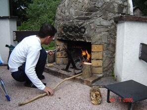 Een buitenhaard van natuursteen met een brandend vuur, een houten aanmaakhout op de grond, en een metalen tafel in de buurt.