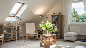 Una sala de estar moderna con techo inclinado, un telescopio, un mueble de madera, una silla, una mesa de centro de vidrio y vistas a árboles a través de un ventanal grande.