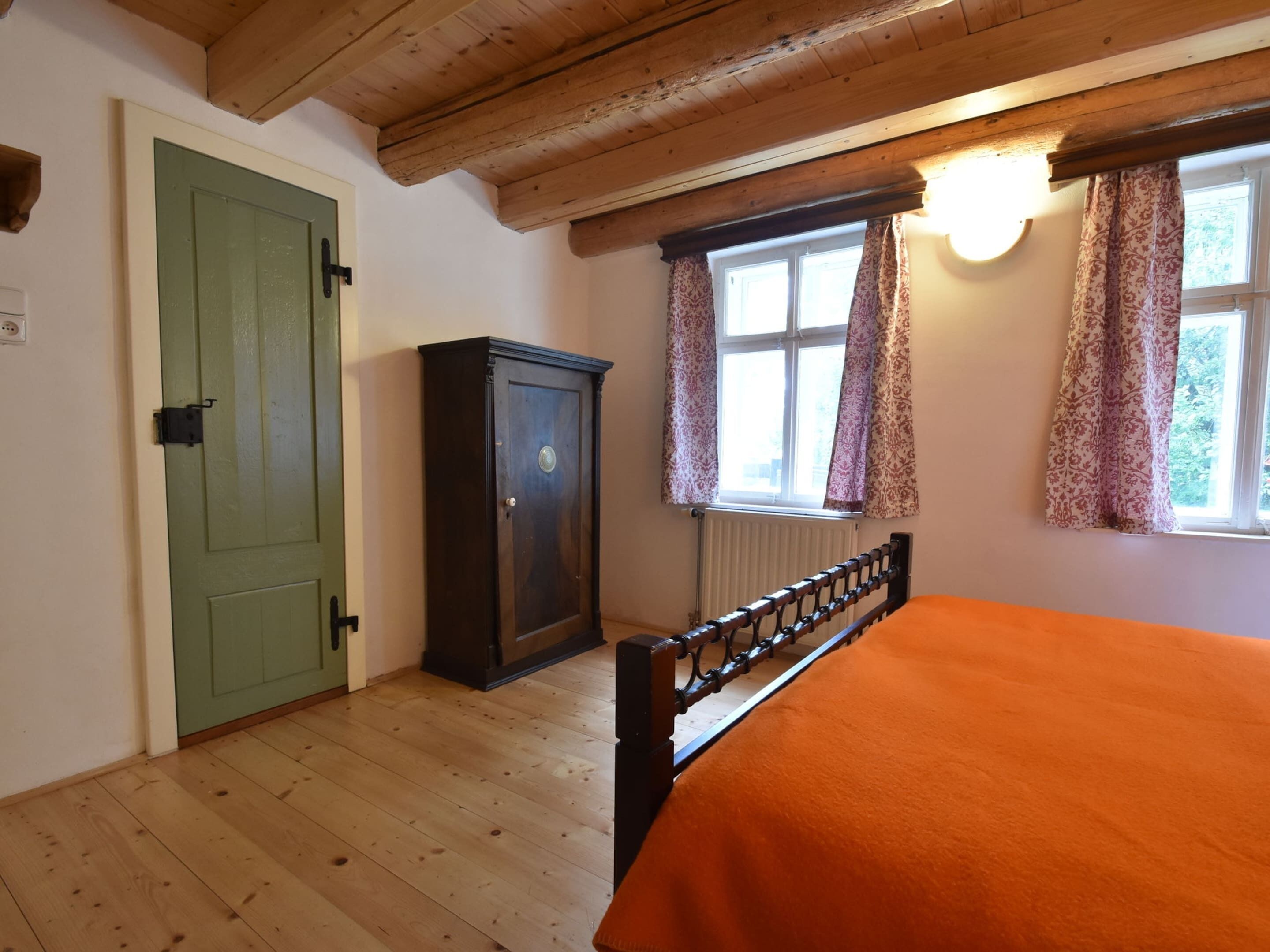 A bedroom with a wooden floor, a green door, a dark wooden cabinet, a bed with an orange bedspread, and a window with patterned curtains.