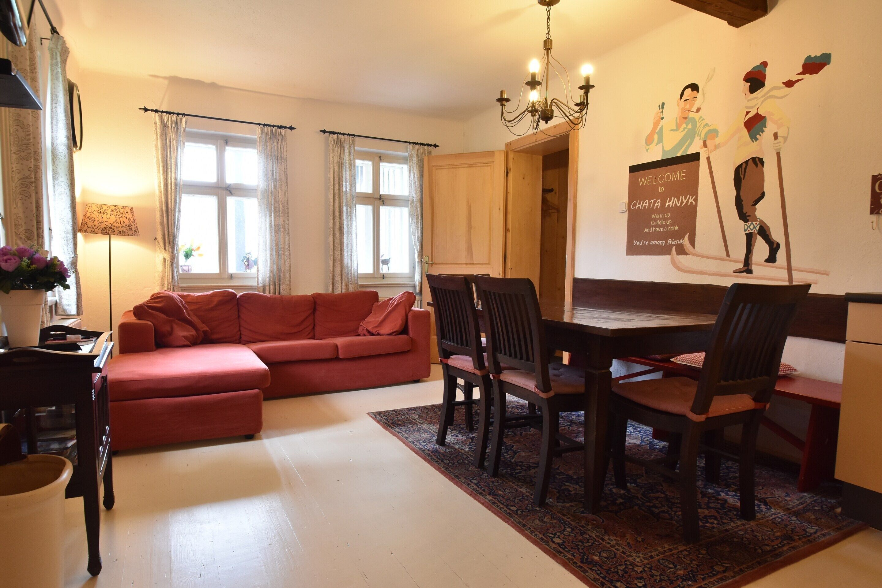 A living room with a red sofa, wooden dining table, and chairs, a chandelier, and a welcome sign on the wall.