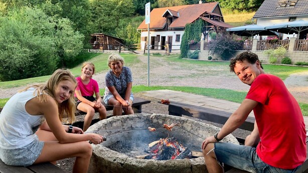 A group of people around a campfire in a backyard with a house and a wooden deck in the background.