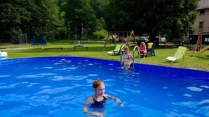 A swimming pool with a clear blue water surface, surrounded by a grassy area with trees and a playground in the background.
