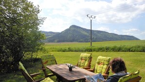 A person sitting at a wooden table outdoors with a book, surrounded by chairs with cushions.