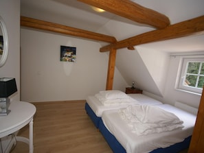 A bedroom with two beds, a small table, a lamp, a painting on the wall, and a window.