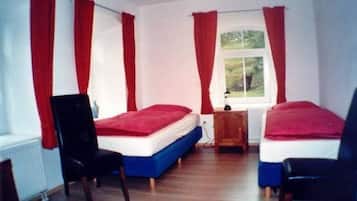 A room with two single beds, each with a red blanket, wooden chairs, and a window with red curtains.