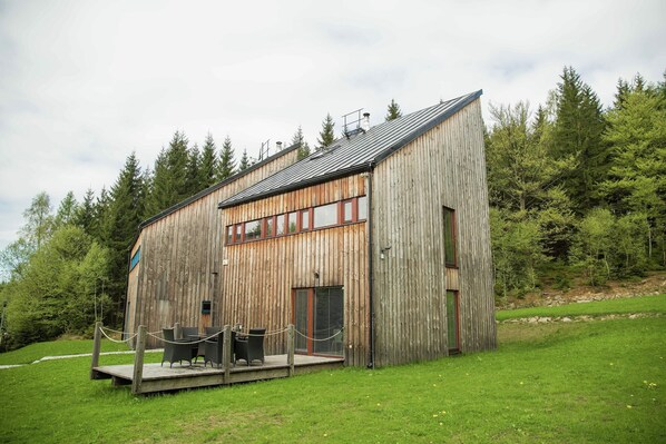 A rustic wooden cabin with a deck, surrounded by a forest.