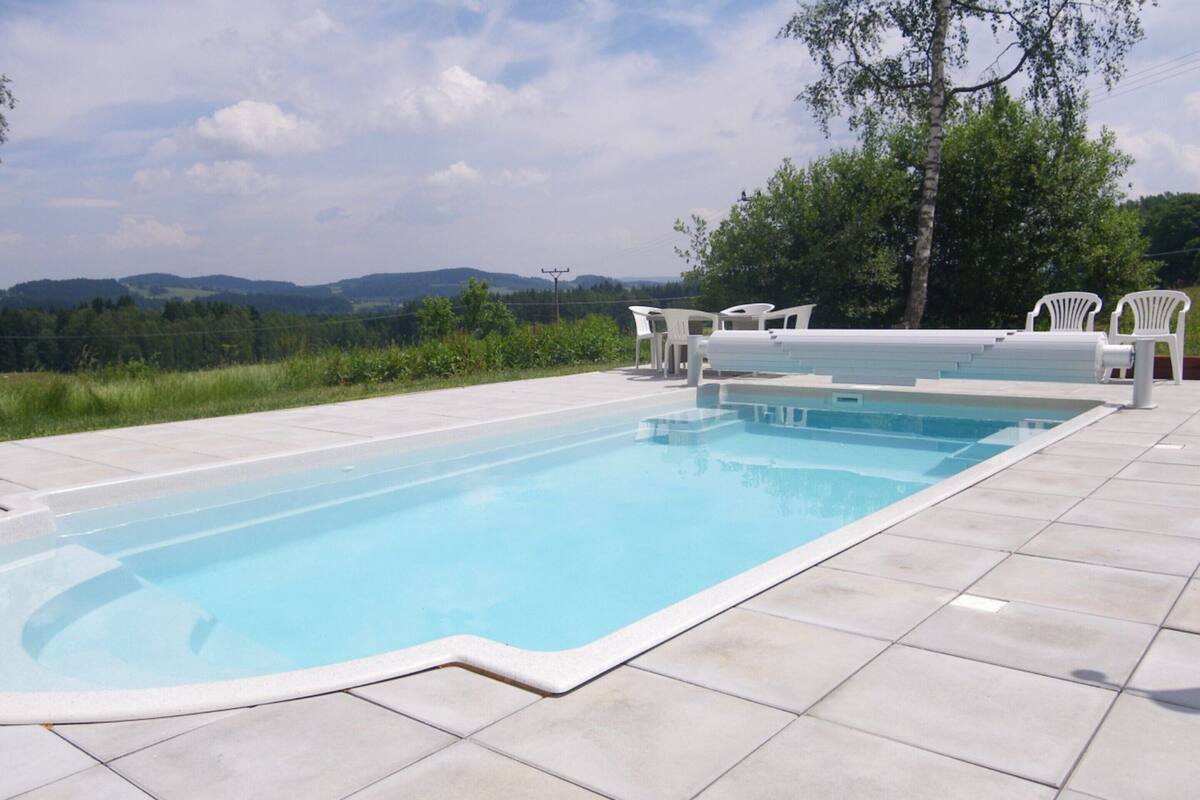 Una piscina rectangular con un borde empedrado, rodeada de sillas blancas y un árbol.